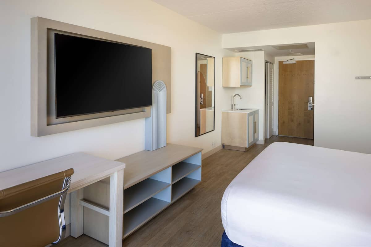 Ein Hotelzimmer mit einem Bett, einem an der Wand befestigten Fernseher, einem Schreibtisch mit Stuhl, einem Spiegel und einem Badezimmer mit Waschbecken und Toilette.