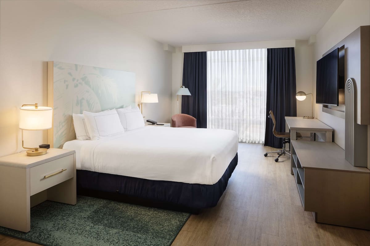Ein Hotelzimmer mit Bett, Schreibtisch, Stuhl, Fernseher und einem großen Fenster mit Vorhängen.