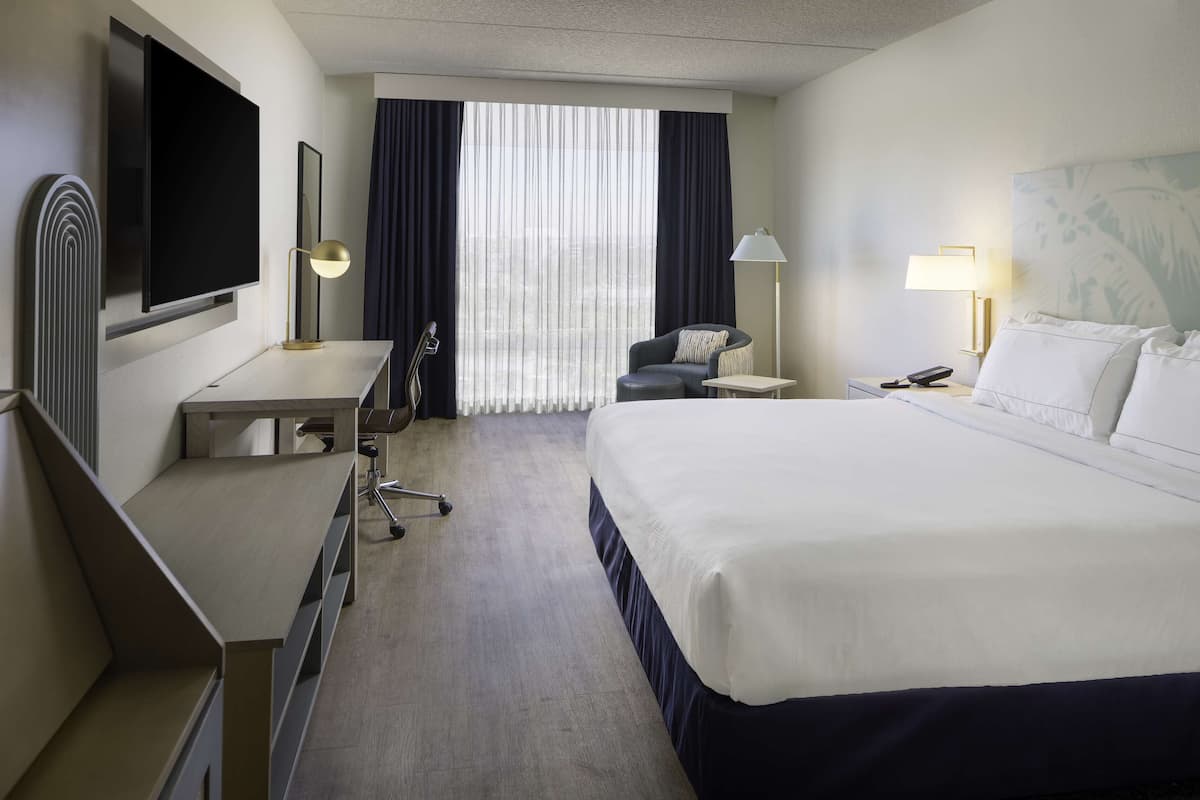 Ein Hotelzimmer mit Bett, Schreibtisch, Stuhl, Fernseher und einem Fenster mit Vorhängen.