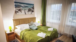 Una cama bien hecha con una colcha verde y almohadas blancas, una mesita de noche con una lámpara y un cuadro de una escena de playa en la pared.