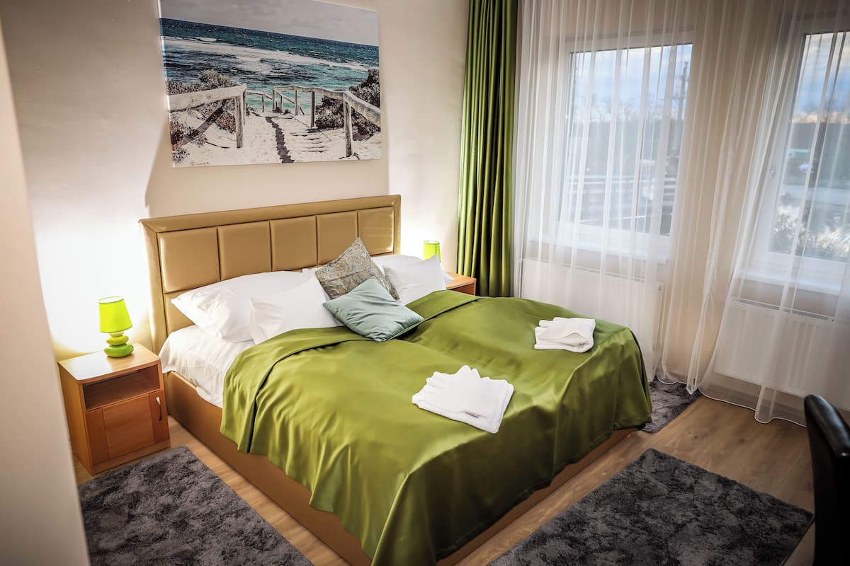 Ein ordentlich bezogenes Bett mit grüner Decke und weißen Kissen, ein Nachttisch mit Lampe sowie ein Bild mit Strandmotiv an der Wand.