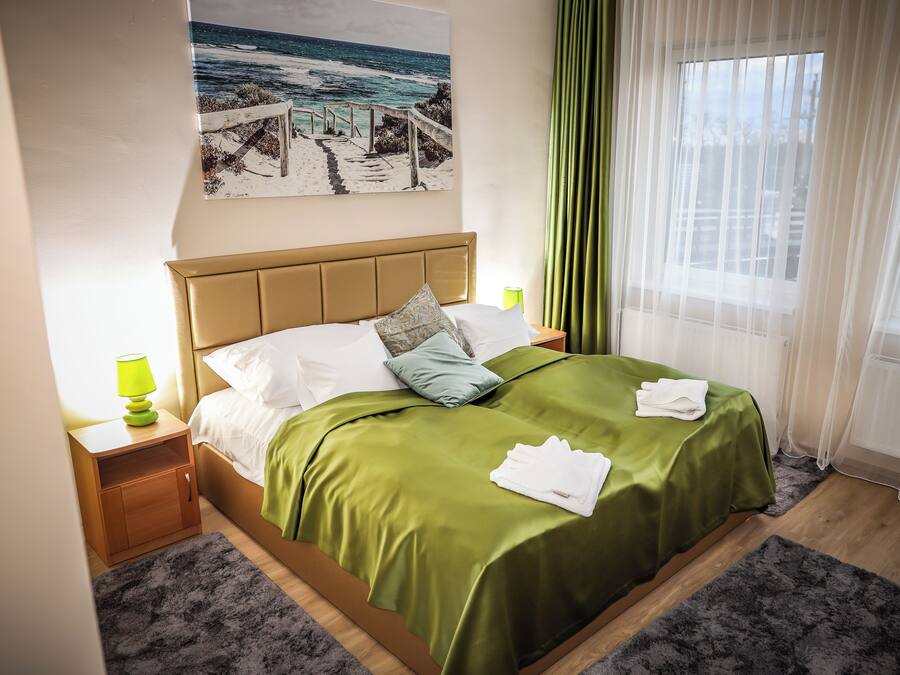 Ein ordentlich bezogenes Bett mit grüner Decke und weißen Kissen, ein Nachttisch mit Lampe sowie ein Bild mit Strandmotiv an der Wand.