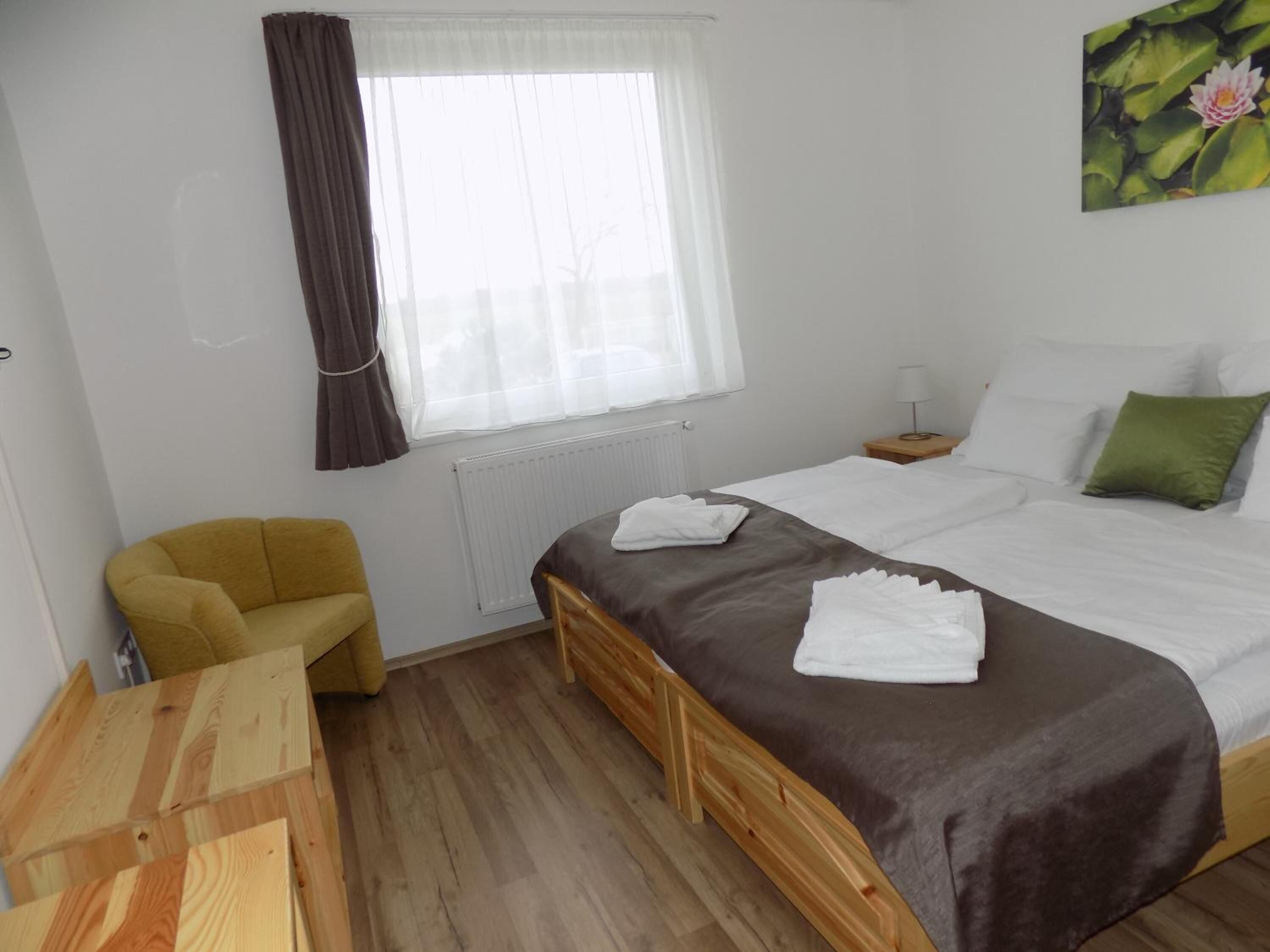 A bedroom with a bed, a chair, a wooden table, a window with curtains, and a painting on the wall.