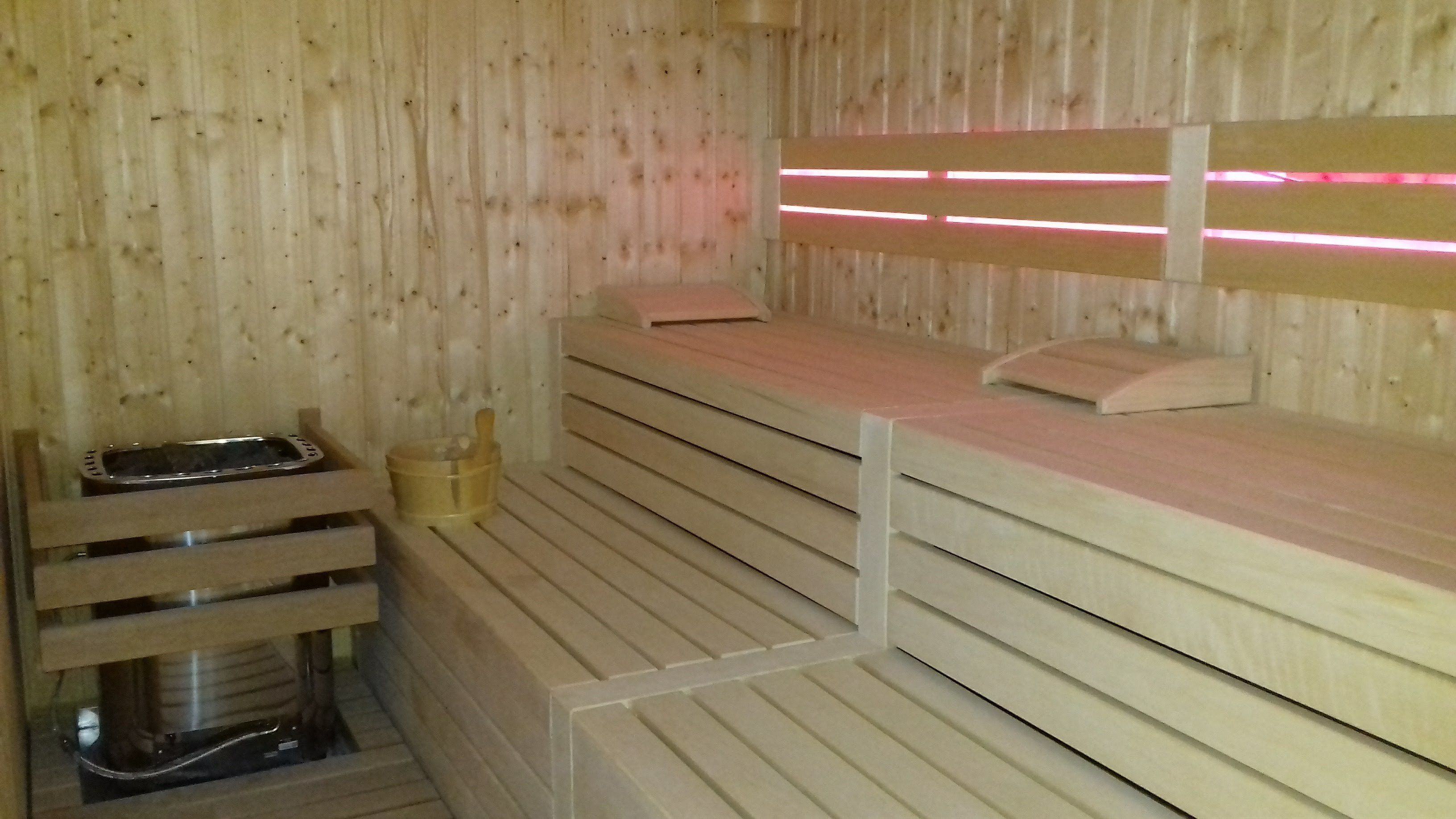 A sauna room with wooden benches, a bench heater, and a wooden wall with a pink light strip.