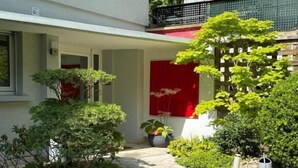 A pathway leading to a building with a red door, surrounded by greenery and a stone-paved area. - Le Jardin de la Tuilerie (Pfastatt)