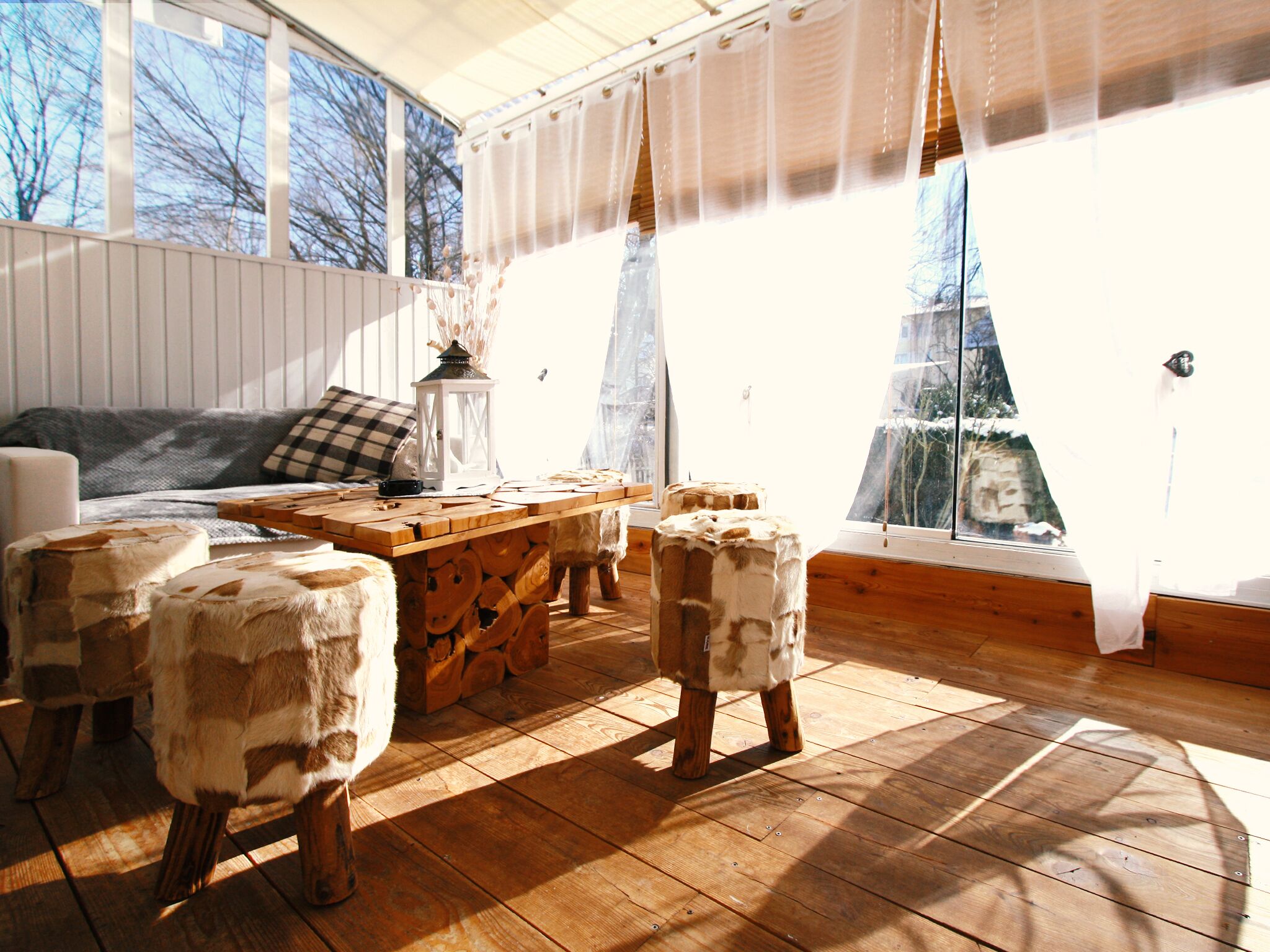 Une pièce chaleureuse avec une table en bois, deux tabourets, un canapé et une fenêtre donnant sur des arbres.