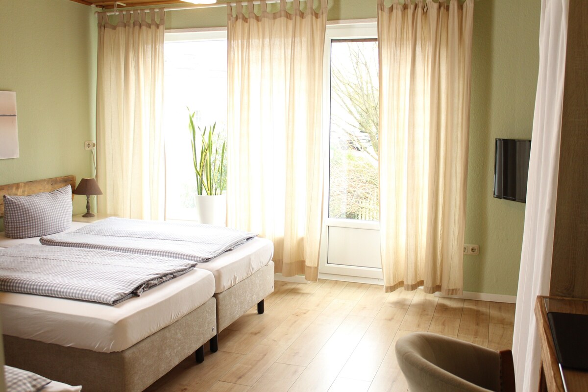 A bedroom with a bed, bedside table, lamp, and a view of the outdoors through a window with curtains.