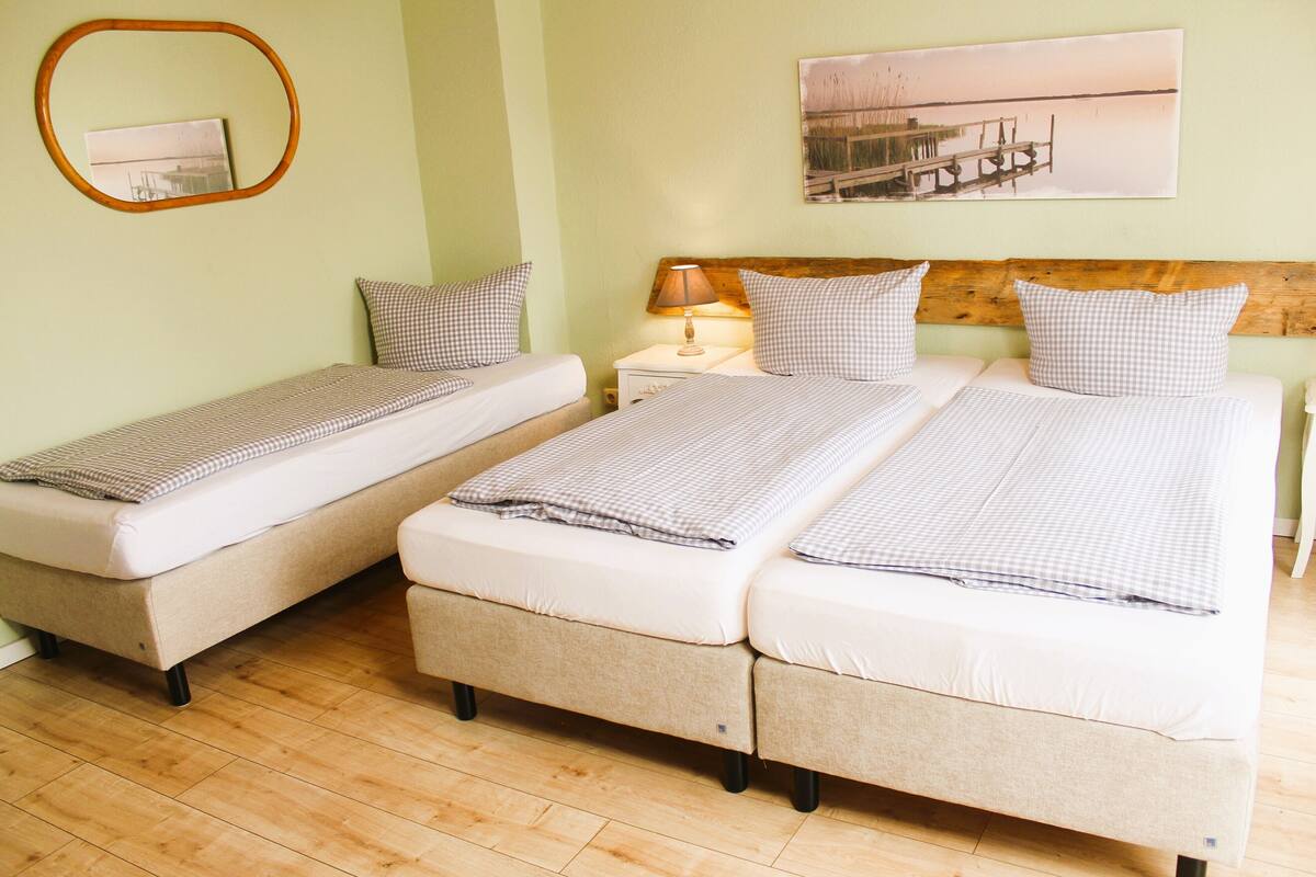 Ein Zimmer mit zwei Einzelbetten und einem Doppelbett, Holzboden, einem Nachttisch mit Lampe, einem gerahmten Bild an der Wand und einem Spiegel.