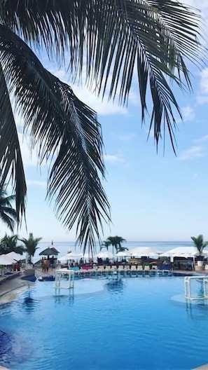 A pool area with clear blue water, surrounded by palm trees and umbrellas, with a view of the ocean in the background.