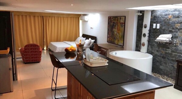 A modern hotel room with a large bed, a sitting area with a table and chairs, a bathtub, and a stone wall. - Résidence Ecologe (Abidjan)