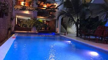 An indoor swimming pool with a blue hue, surrounded by plants and wooden seating.