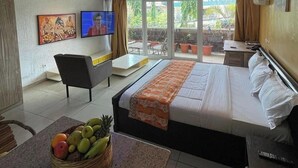 Una moderna camera d'albergo con un letto grande, una TV, un balcone con piante in vaso e un cesto di frutta sul pavimento.