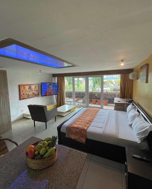 Una habitación de hotel moderna con una cama grande, un televisor, un balcón con plantas en macetas y una cesta de frutas en el suelo.