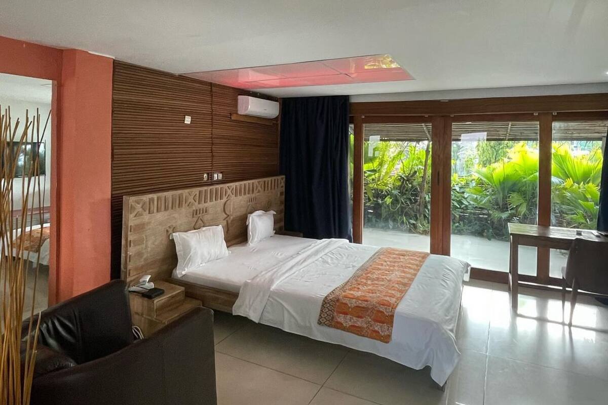 A modern hotel room with a large bed, a sofa, and a view of greenery outside.