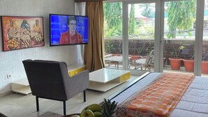 Camera d'albergo con un letto, una televisione, una scrivania con un cesto di frutta, una sedia e un balcone con piante in vaso.