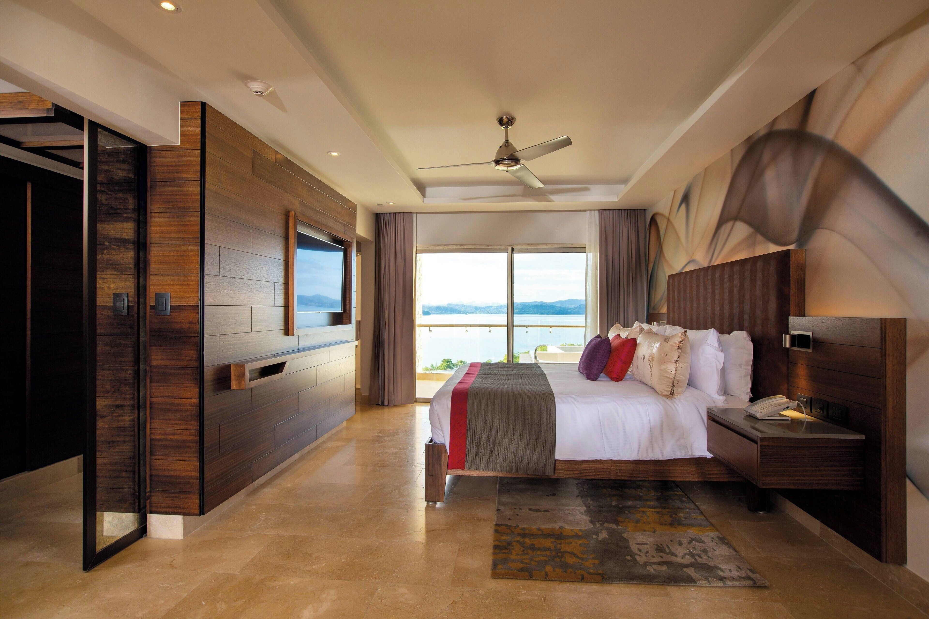 A modern hotel room with a large bed, a ceiling fan, a TV, and a view of the mountains.