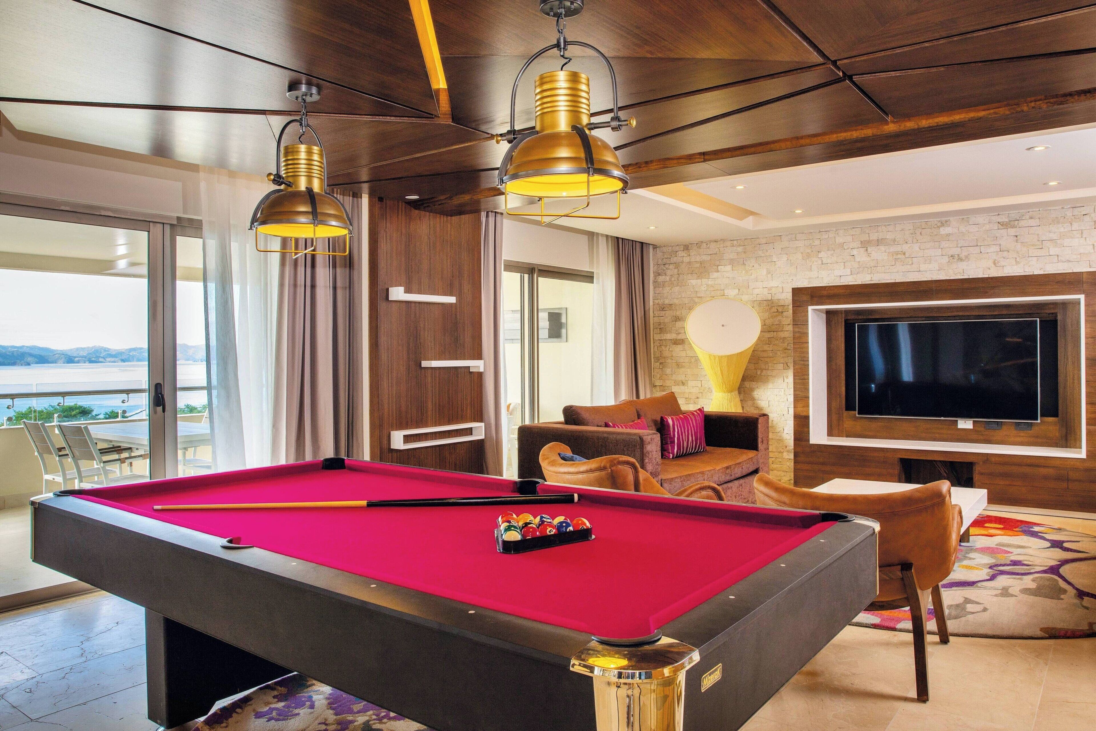 A pool table with a red felt surface, a pool cue, and a rack of balls. There is a television mounted on the wall and a sofa with cushions. The room has a wooden ceiling and a brick wall.
