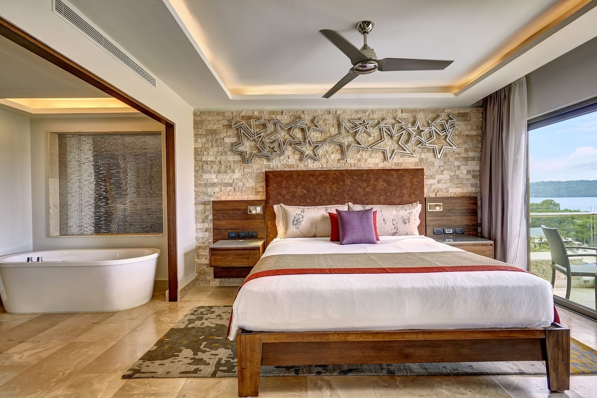 A modern bedroom with a large bed, a ceiling fan, a stone wall with star decorations, a bathtub, and a view of the outdoors.