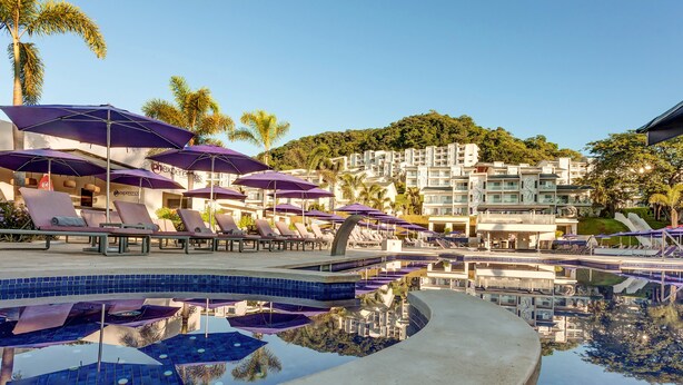 A pool area with lounge chairs and umbrellas, a reflection pool, and a view of hotel buildings with balconies. - Planet Hollywood Costa Rica by Royalton, An Autograph Collection All-Inclusive Resort (Nacascolo)