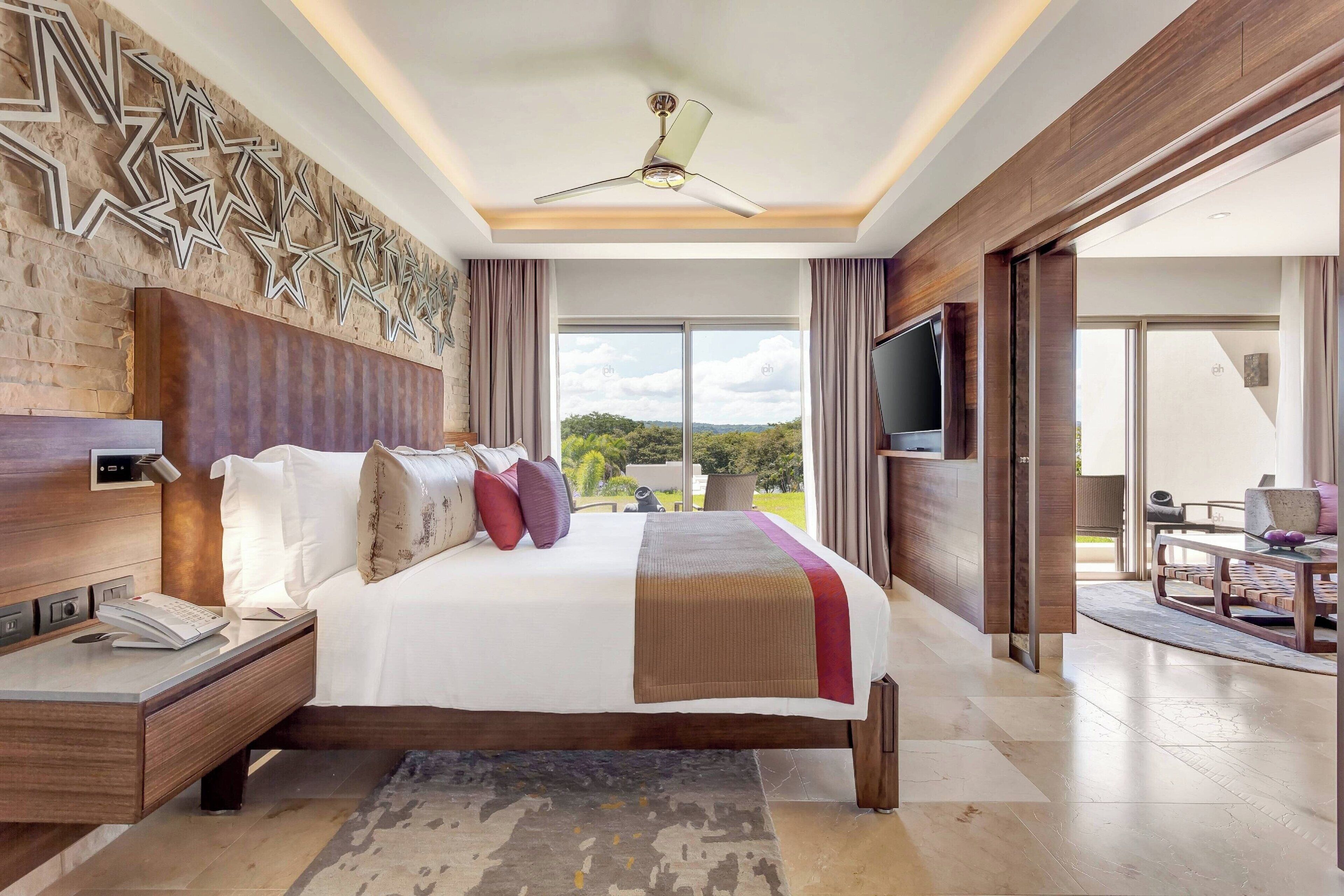Een moderne hotelkamer met een groot bed, een flatscreen televisie, een plafondventilator en uitzicht op de omgeving.