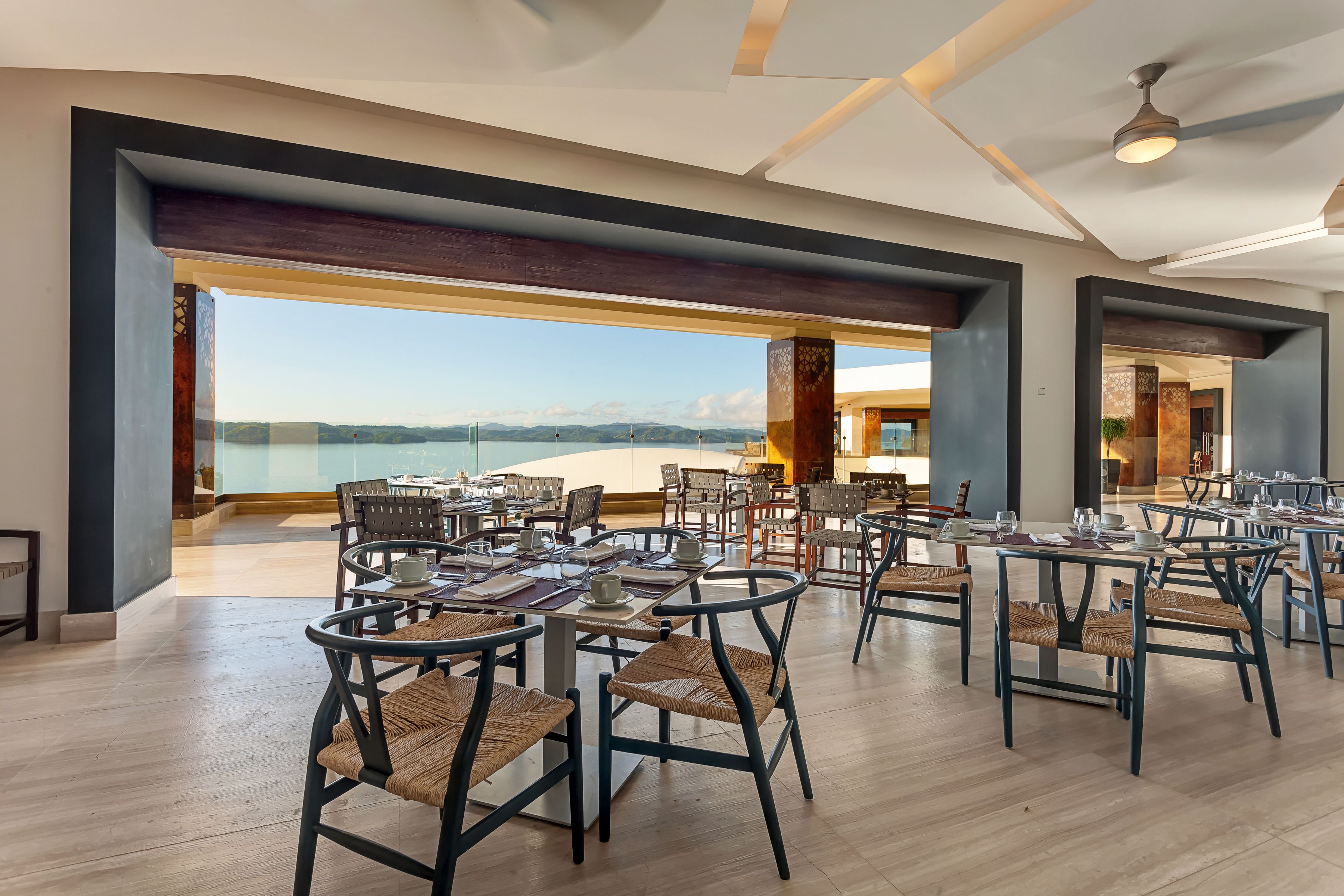 Una àmplia zona de dinar amb taules i cadires, grans finestres que ofereixen vista al mar i a les muntanyes.
