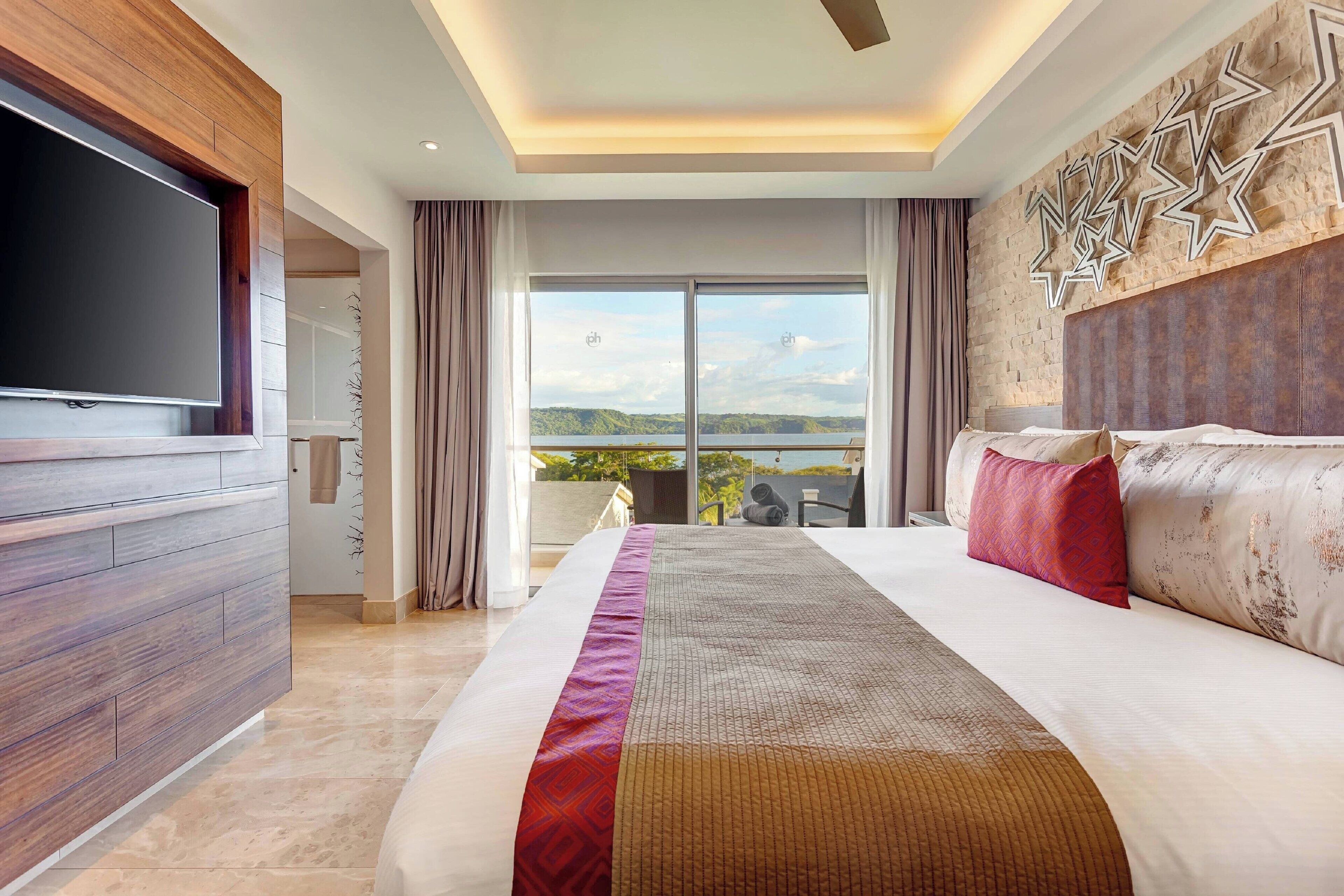 A modern hotel room with a large bed, a flat-screen TV, a balcony with a scenic view, and a decorative wall.