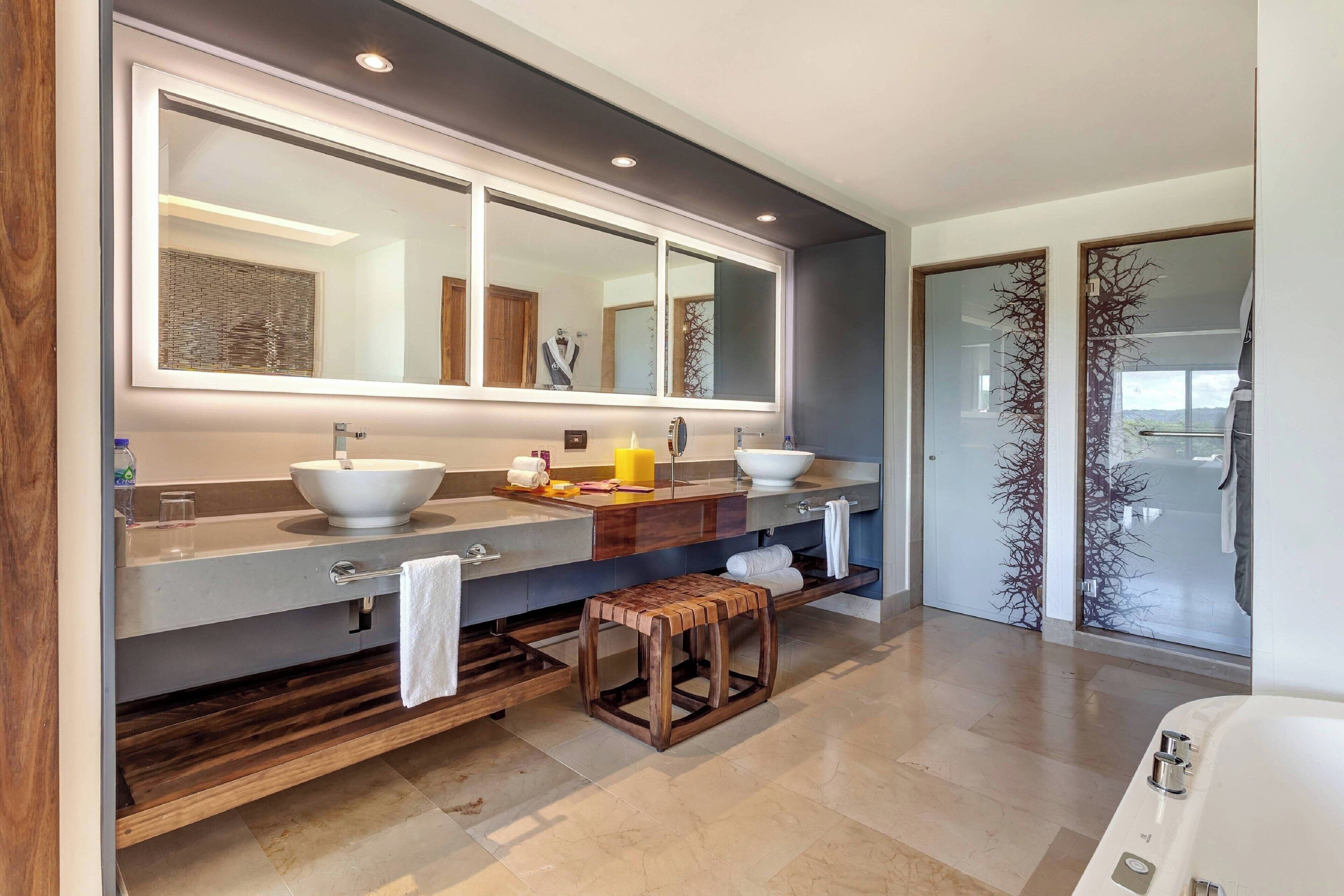 A modern bathroom with a large mirror, two sinks, a wooden stool, and a walk-in shower.