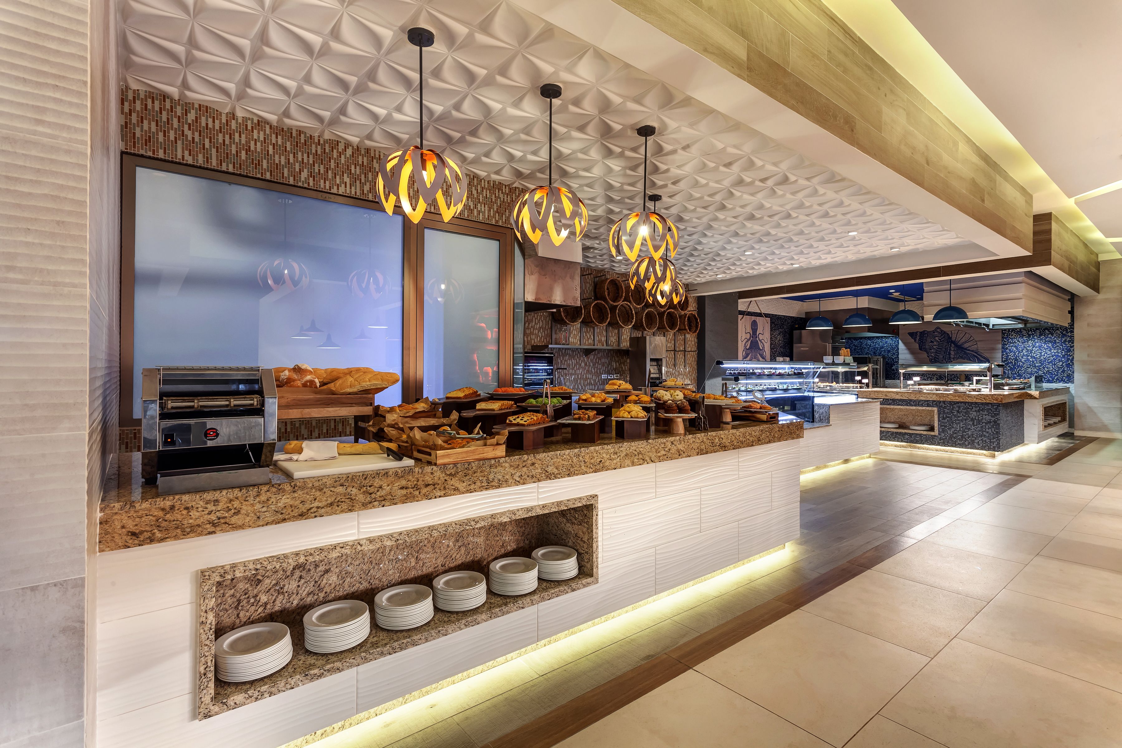 A modern breakfast bar with a built-in coffee machine, a variety of food items, and a seating area with a patterned ceiling.