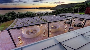 A rooftop terrace with a view of a lake and hills, featuring a seating area with tables and chairs, and a fire pit.