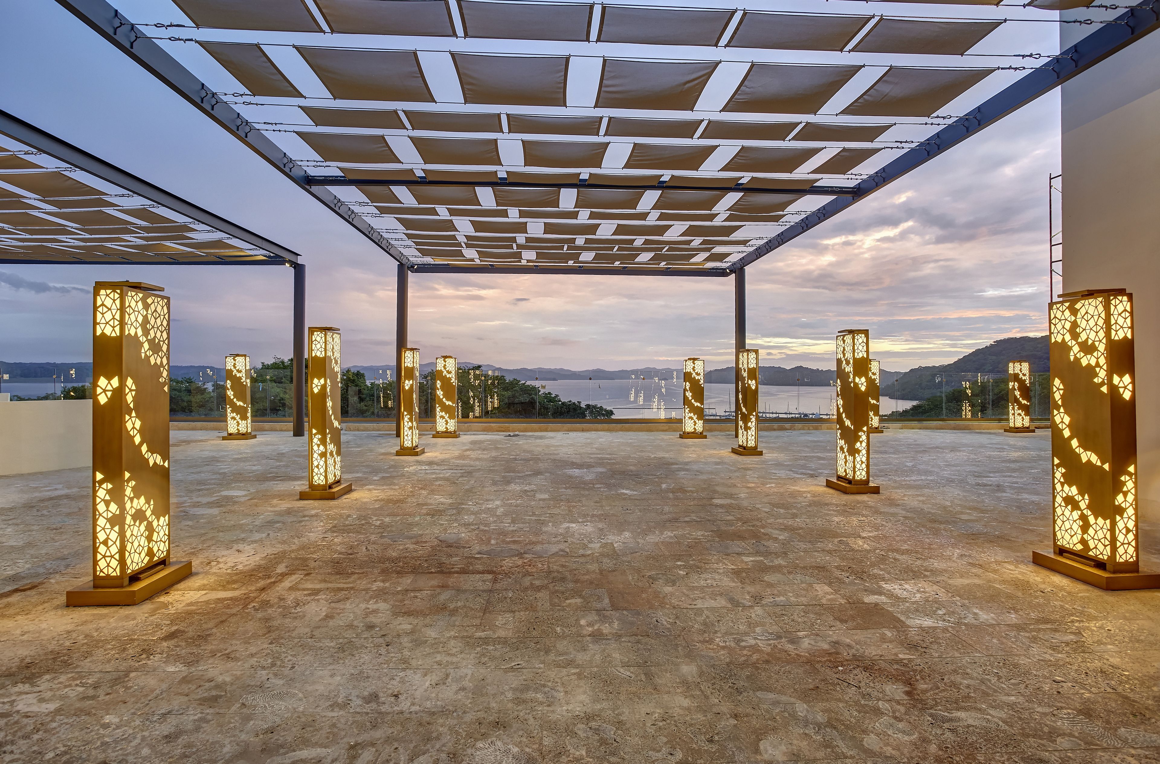Una terraza en la azotea con una pérgola, varios pilares iluminados y vistas a montañas distantes.