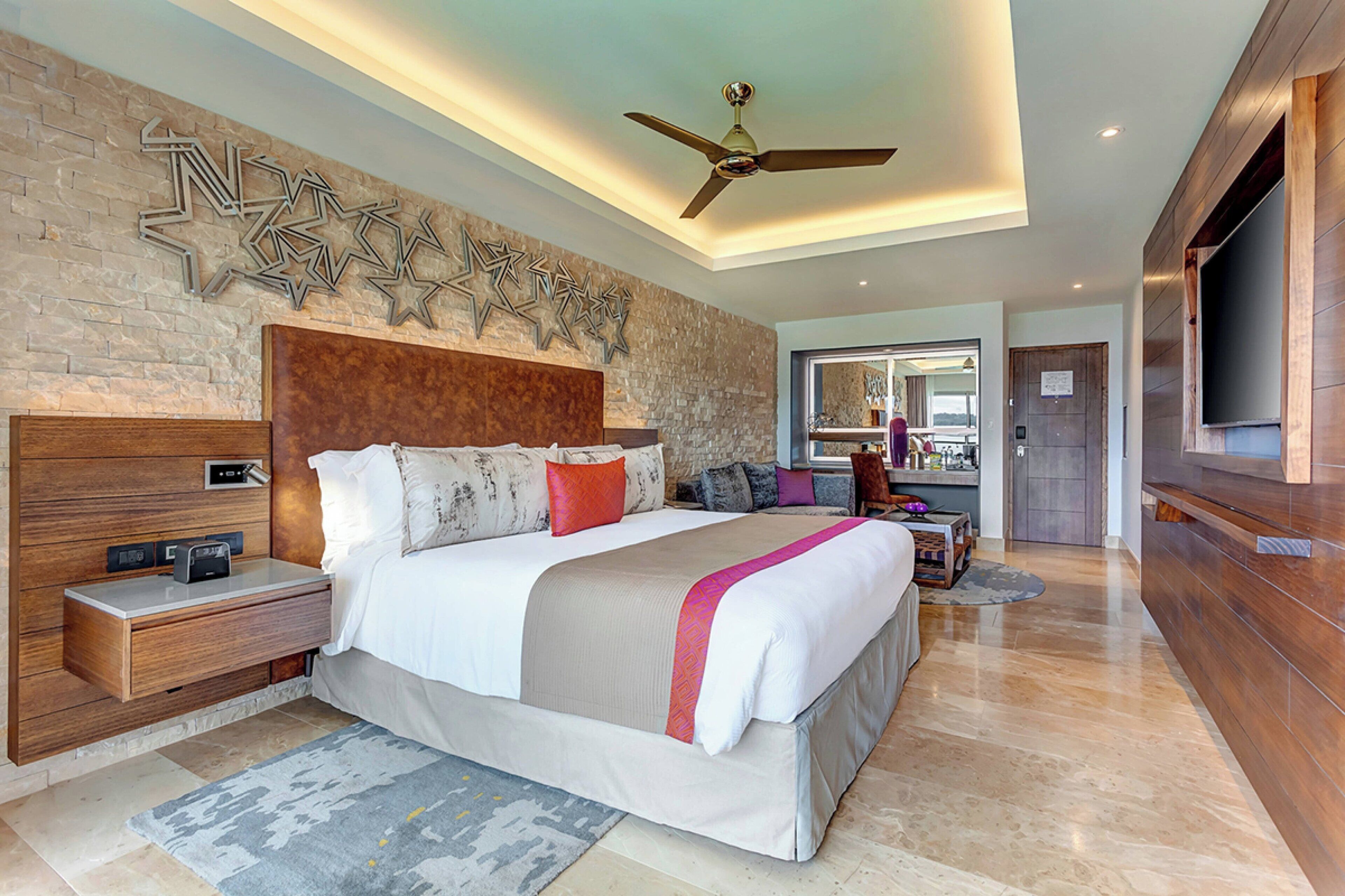 A modern hotel room with a large bed, wooden headboard, a ceiling fan, and a stone accent wall.