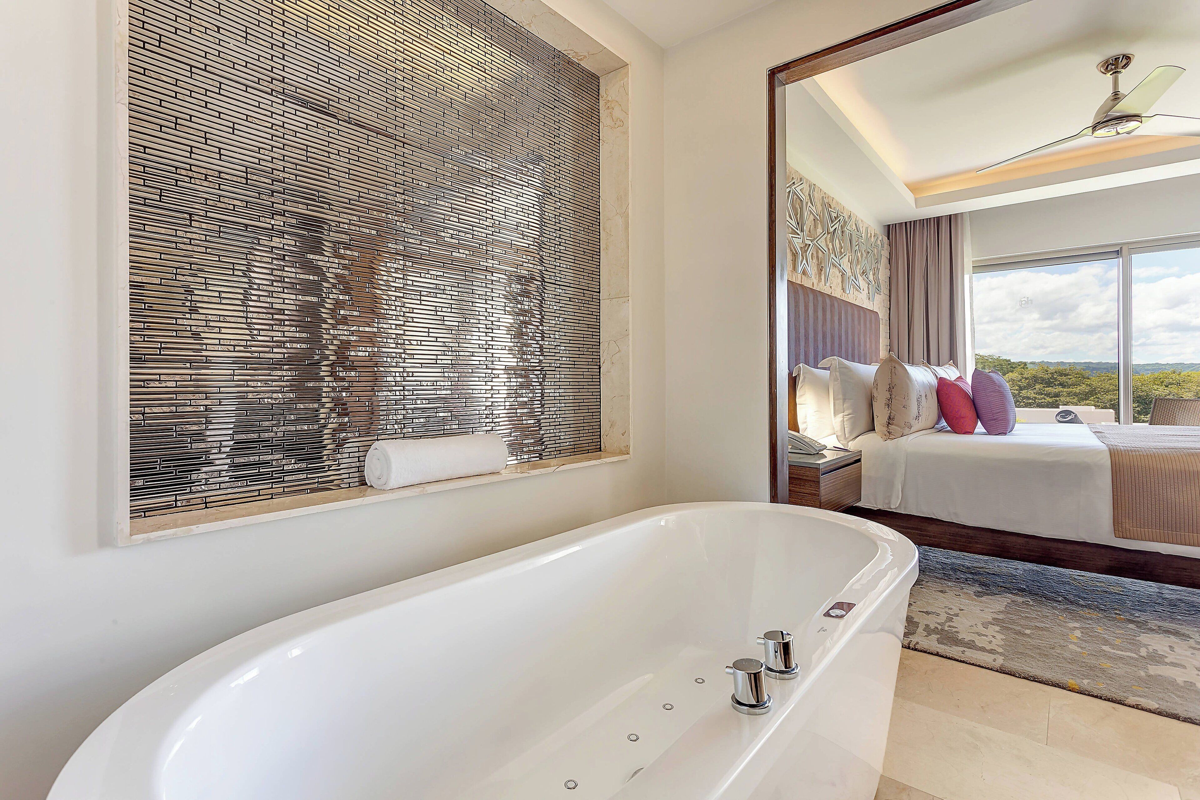 A modern bathroom with a large bathtub, a decorative wall, and a bed with a view.