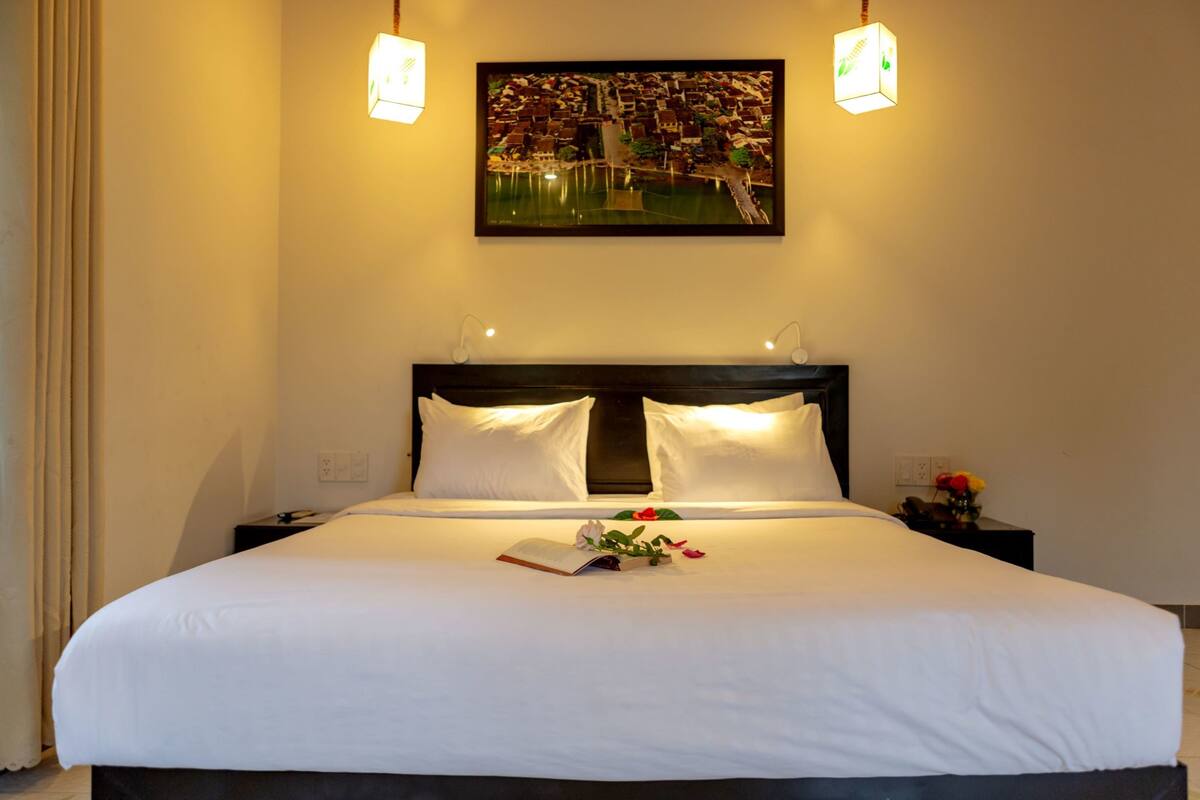 A neatly made bed with white linens, a framed picture above it, and a small table with a vase of flowers beside the bed.