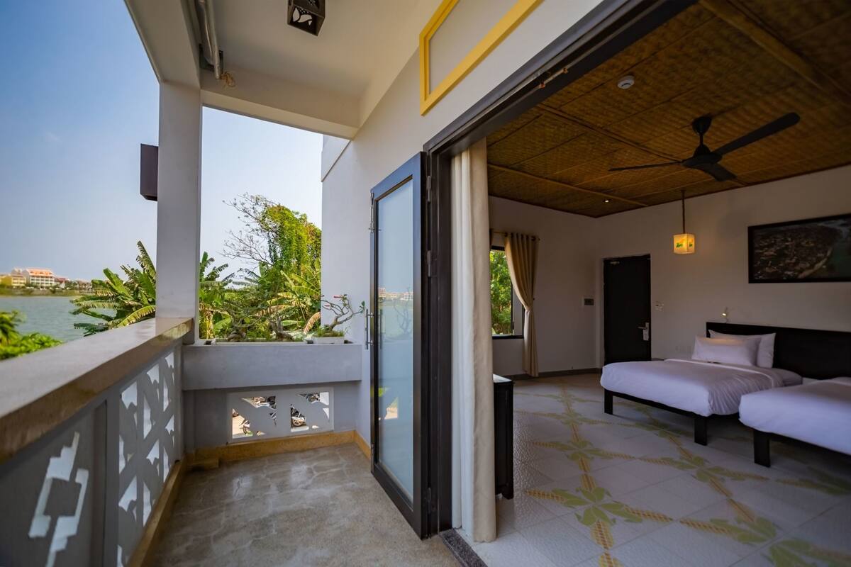 A hotel room with a balcony overlooking a body of water, a bed, a ceiling fan, and a view of greenery.