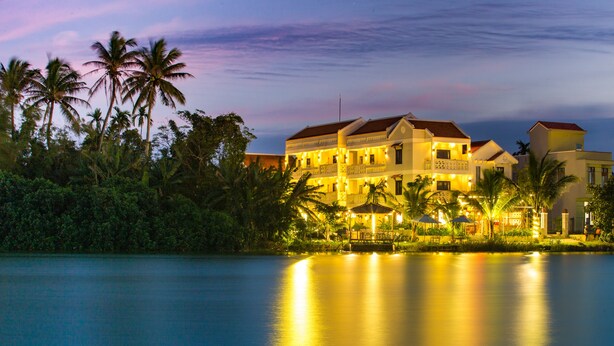 A large, multi-story hotel with a waterfront view, surrounded by palm trees. - Hoi An Corn Riverside Villa (Da Nang)