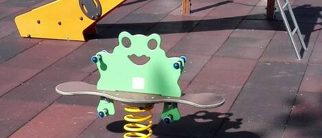 Een speeltuin met een speelconstructie in de vorm van een groene kikker en een gele glijbaan.