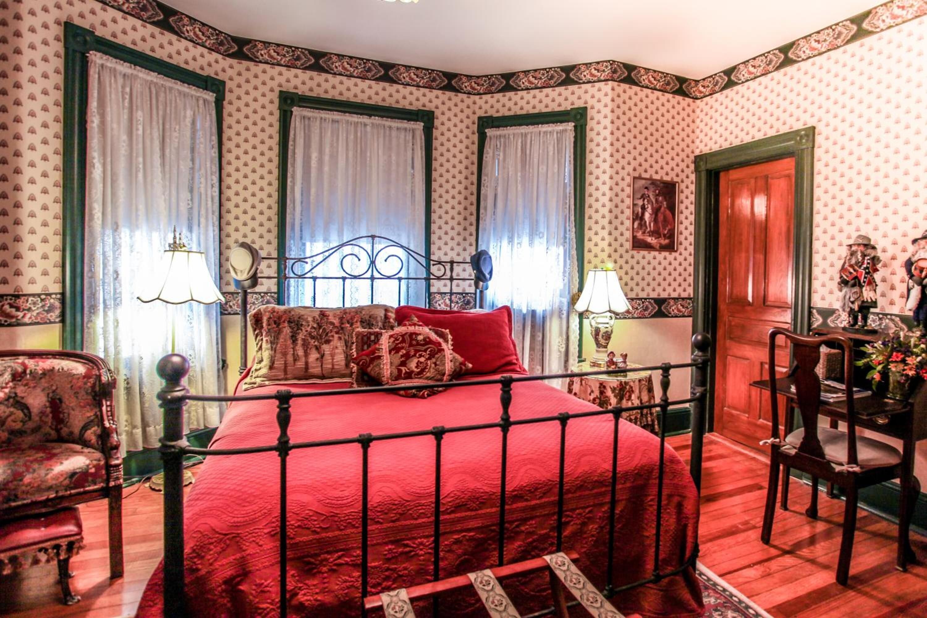A bedroom with a red bedspread, a metal bed frame, two windows with green frames, and a wooden door.