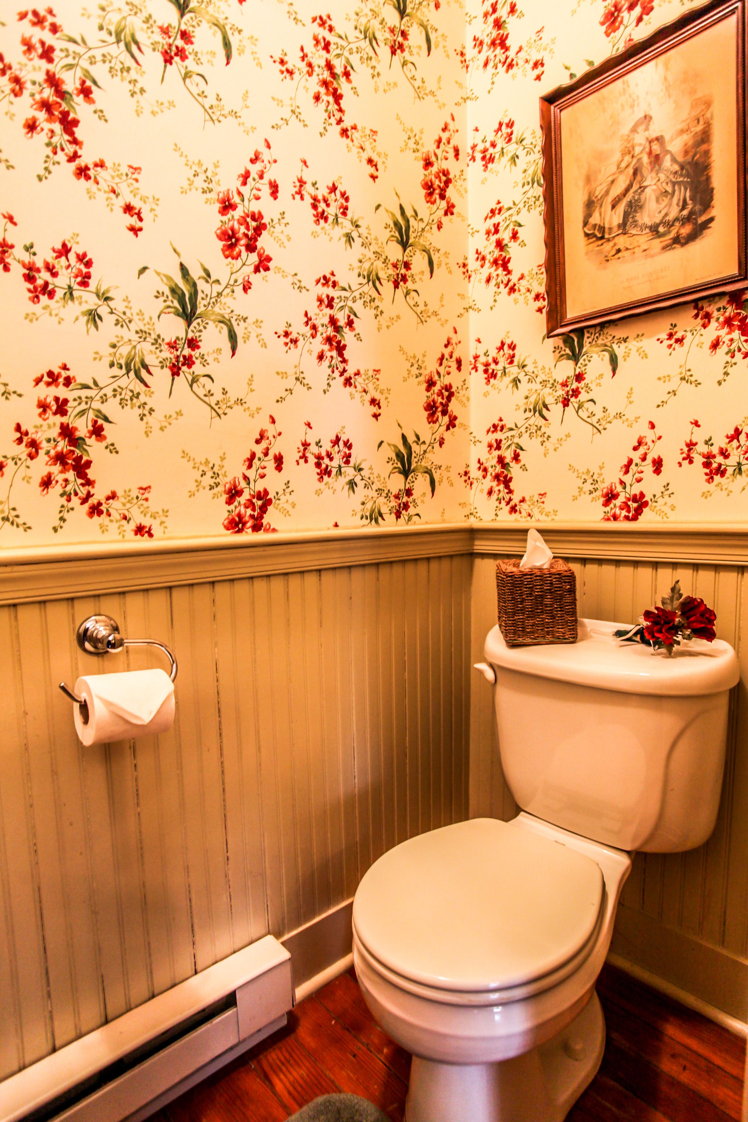 A bathroom with floral wallpaper, a toilet, a framed picture on the wall, and a tissue box on the toilet.
