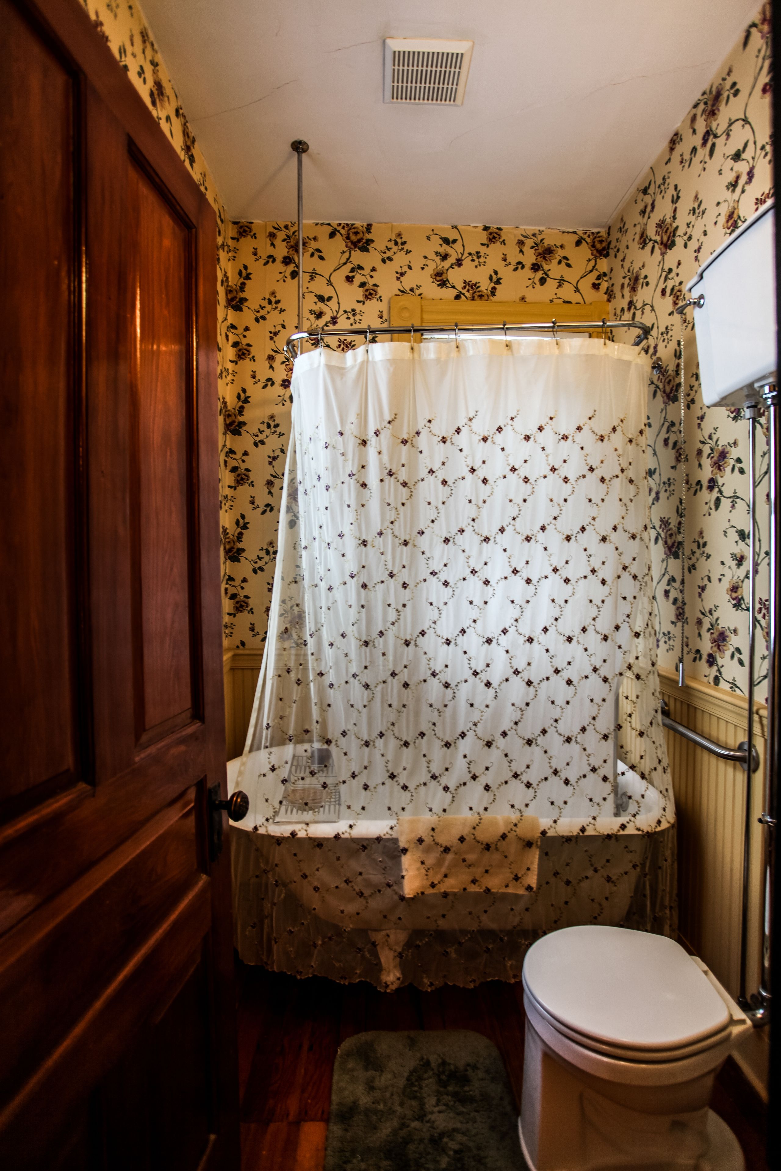A bathroom with a wooden door, a bathtub with a floral shower curtain, and a white toilet.