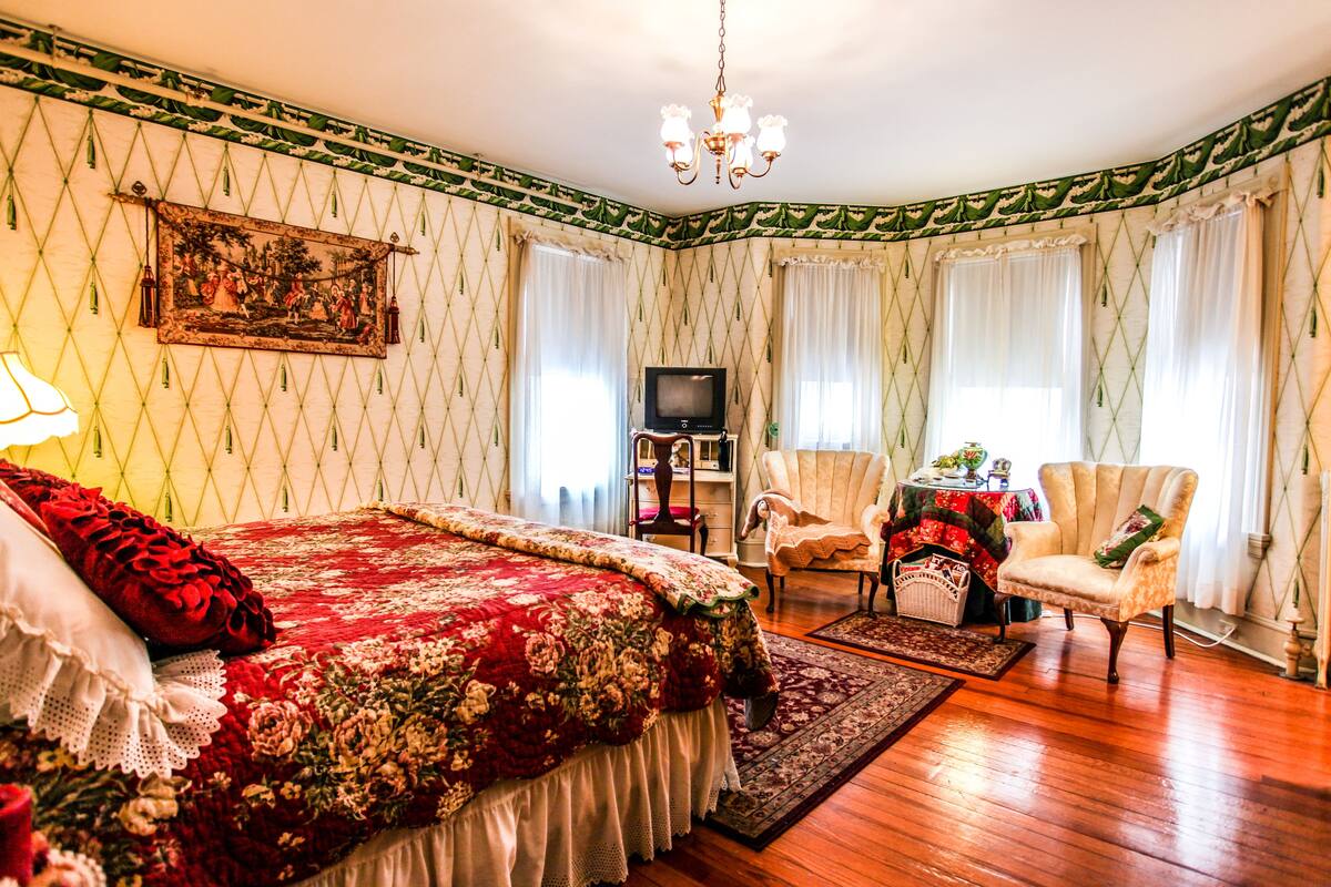 A traditional room with a large bed, a television, a chair, and a table with a floral tablecloth.