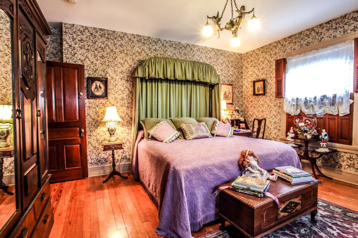 A bedroom with a large bed, wooden floor, floral wallpaper, a green headboard, and a chandelier.