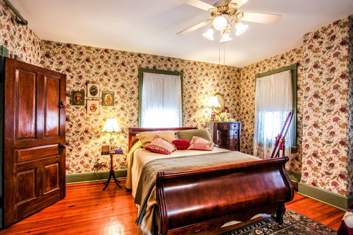 A bedroom with floral wallpaper, a wooden bed, two bedside tables with lamps, a ceiling fan, and a wooden door.