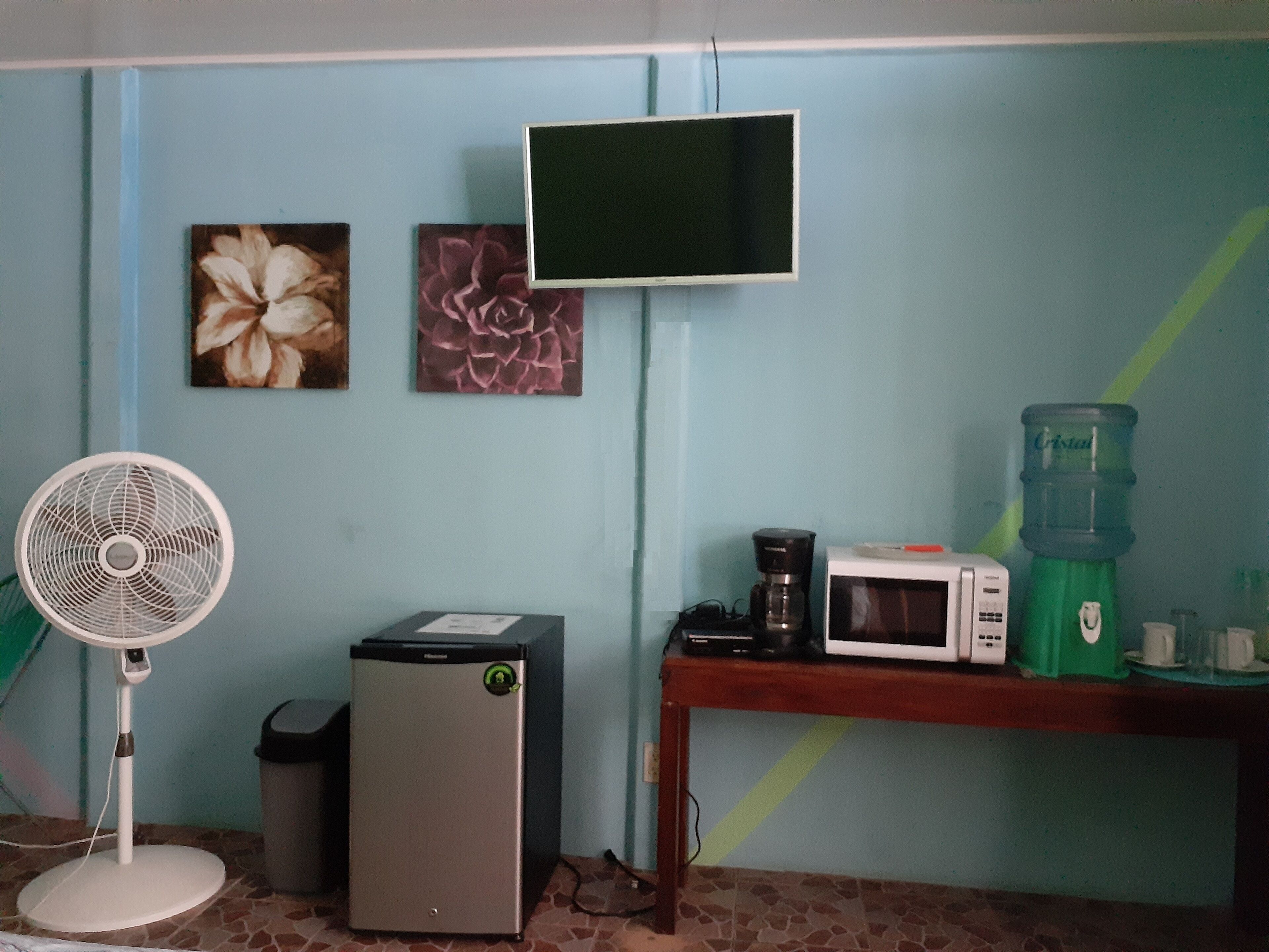 A room with a fan, a small refrigerator, a microwave, and a water dispenser on a table.