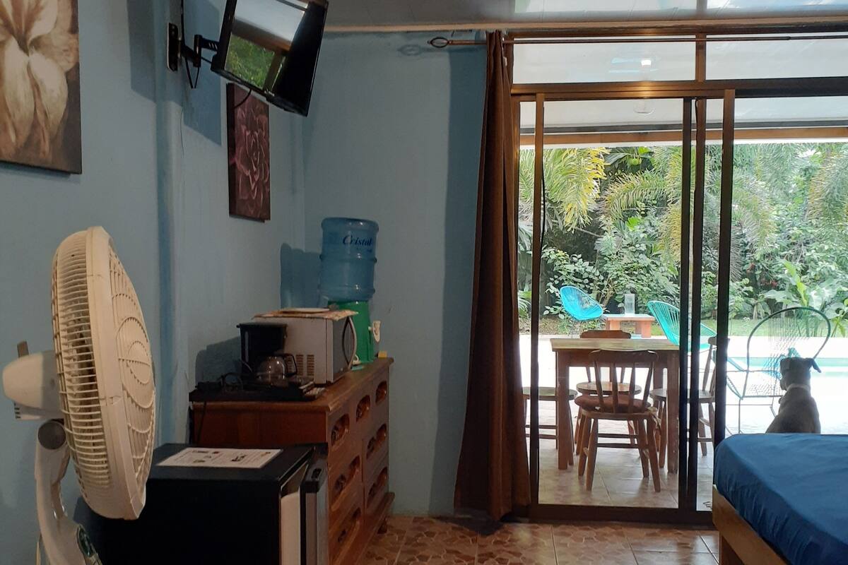 A room with a bed, a desk, a fan, a water dispenser, and a television on the wall.