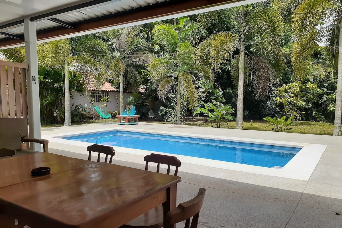 A poolside area with a wooden table and chairs, surrounded by lush greenery and a clear blue pool.