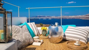 A sunlit terrace with a view of the sea, featuring a wicker sofa, cushions, a lantern, and a decorative vase. - Cape 9 Suites & Villas (Santorini)