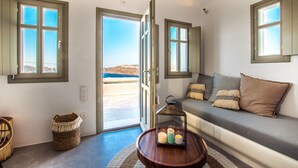 A room with a sofa, a small table with candles, a wicker basket, and a view of the outdoors. - Cape 9 Suites & Villas (Santorini)
