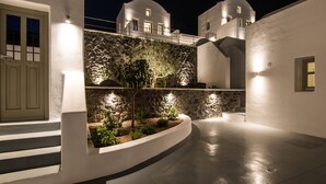 A nighttime view of a white-walled building with stone accents, a curved pathway, and a small garden area. - Cape 9 Suites & Villas (Santorini)