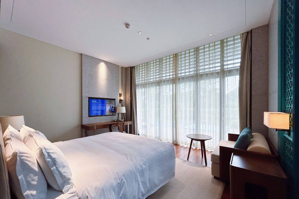 A hotel room with a large bed, a desk with a chair, a TV mounted on the wall, and a window with curtains.