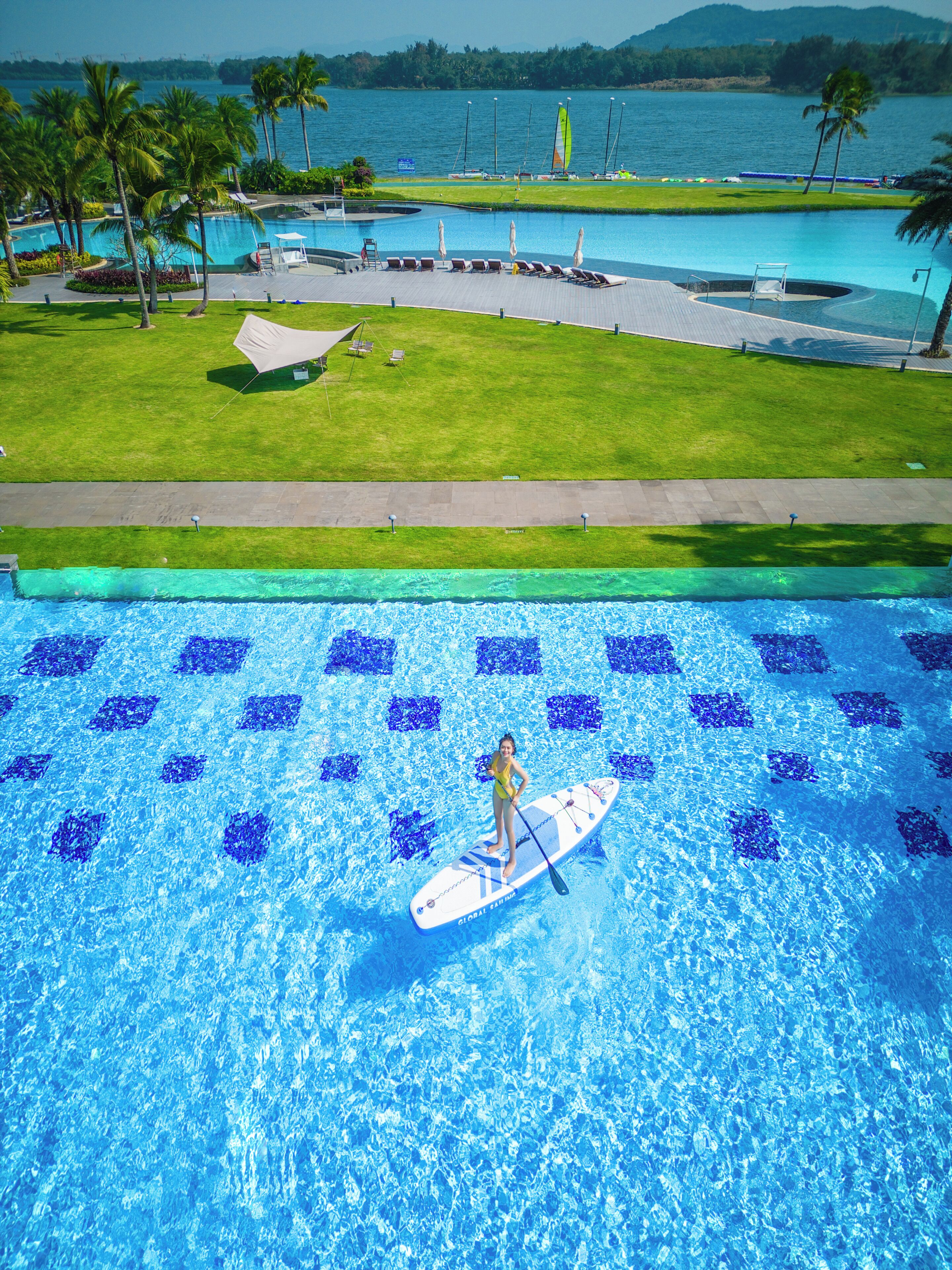 A pool with a person paddleboarding, surrounded by a grassy area and palm trees.